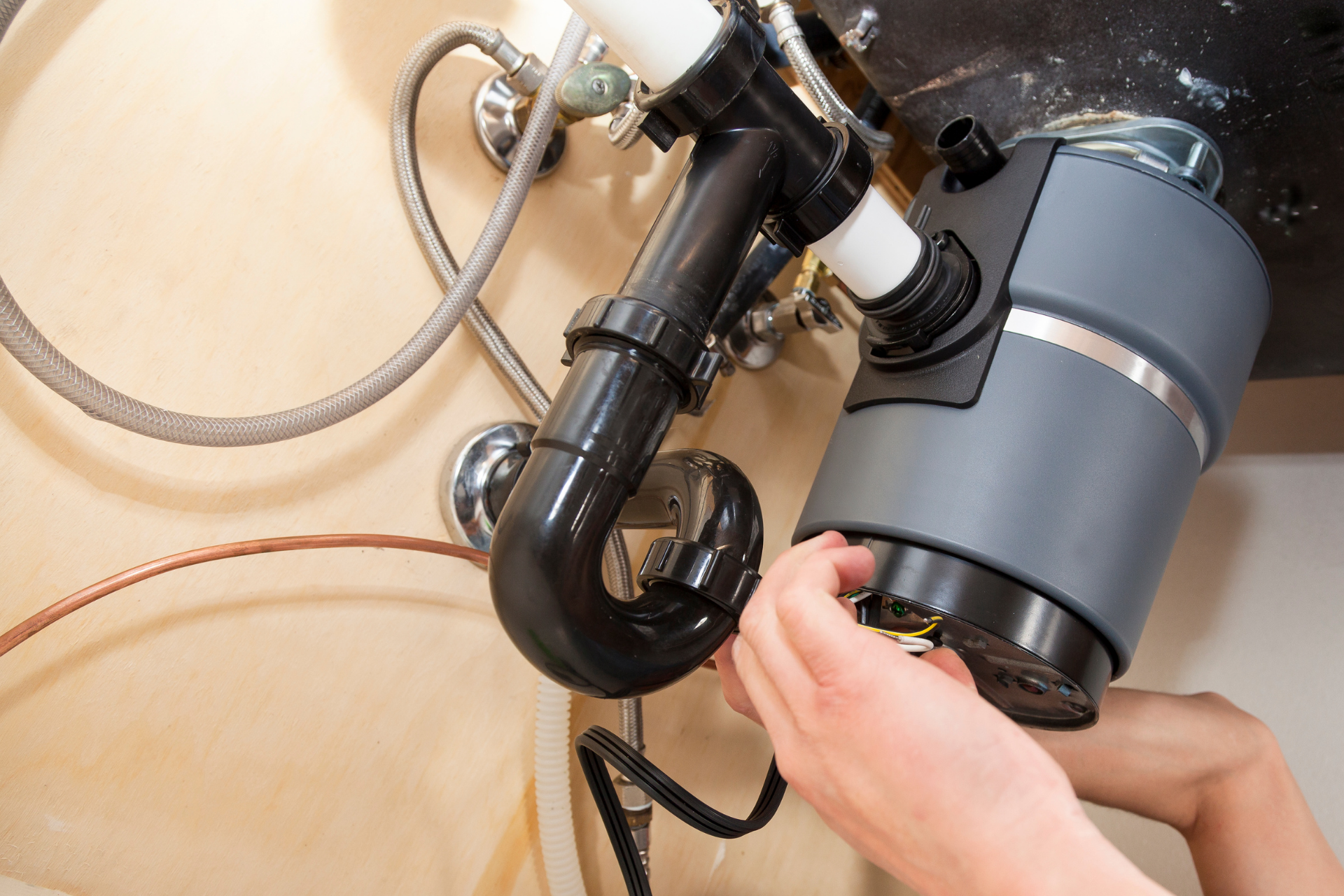 Hands installing a gray garbage disposal under a kitchen sink with exposed pipes