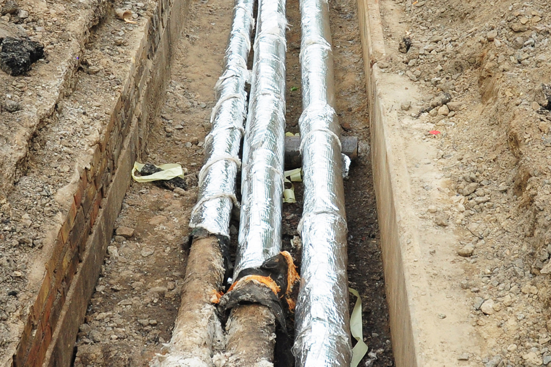 Trenched underground pipes wrapped in silver insulation in a dirt excavation site