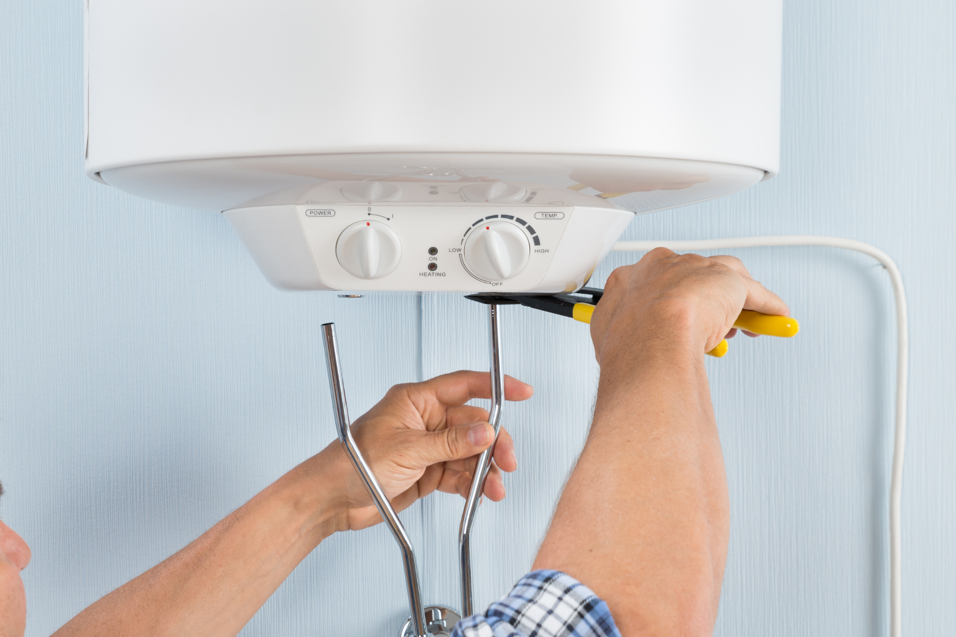 Hands adjusting a white water heater control panel with a screwdriver and hose in a bathroom