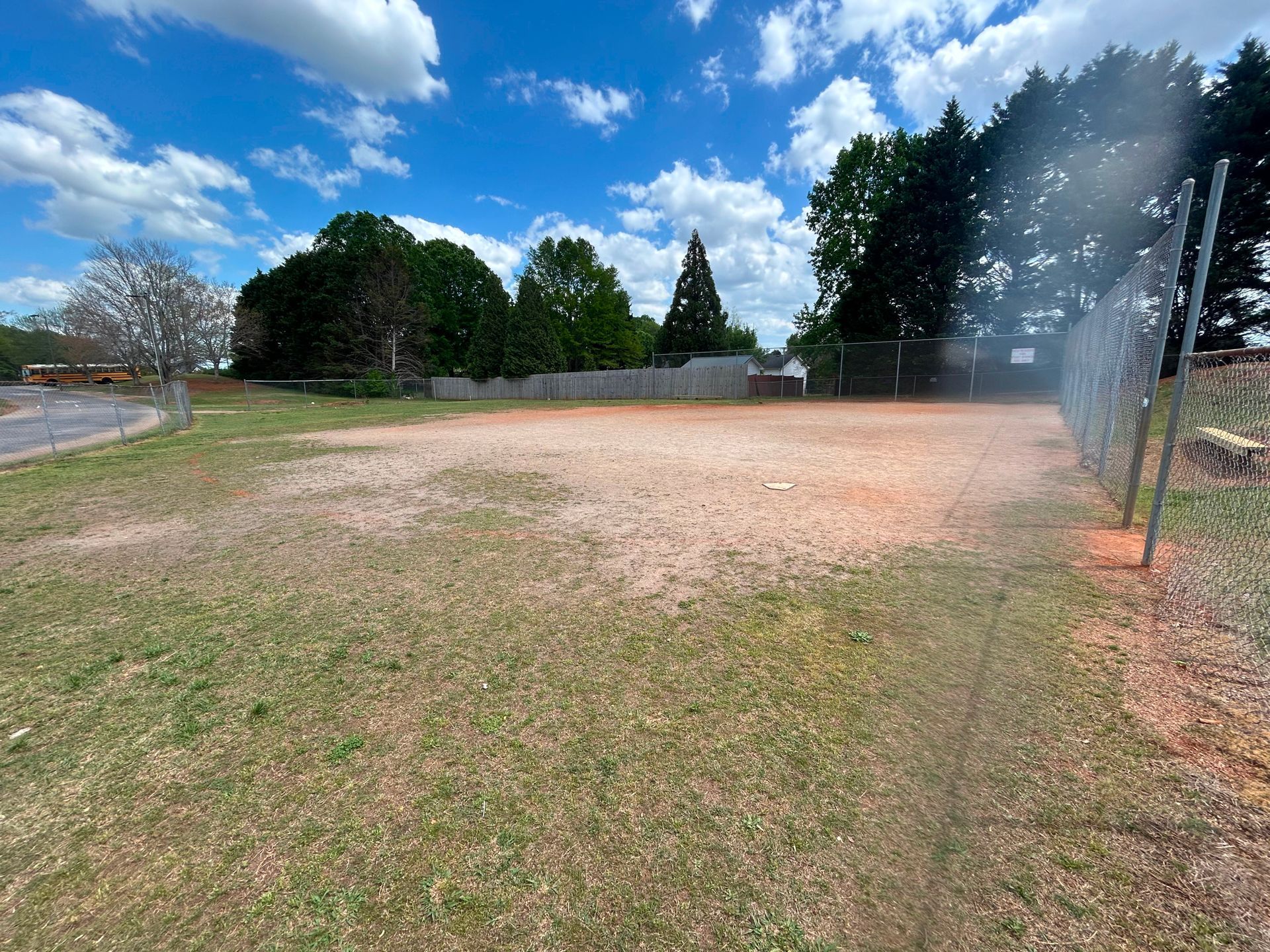 Grassy field with gravel area, surrounded by a chain-link fence and trees under a blue sky.