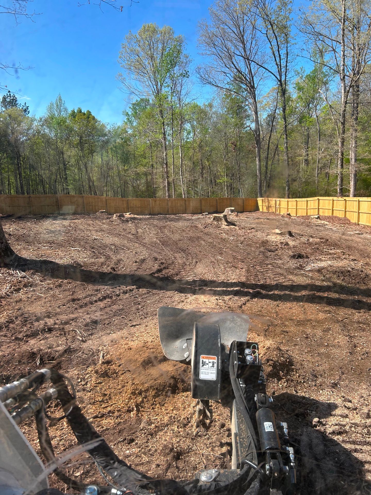 A stump grinder working on a cleared lot with a wooden fence and trees in the background.