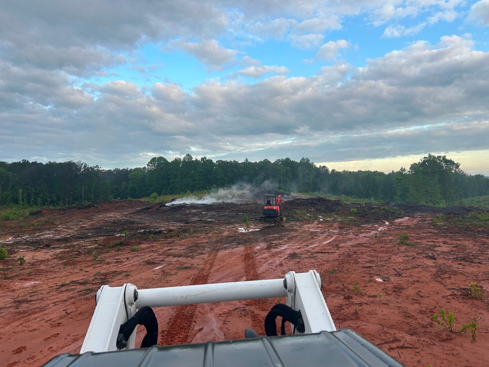 A skid steer driving on muddy ground toward construction equipment, under a cloudy sky.