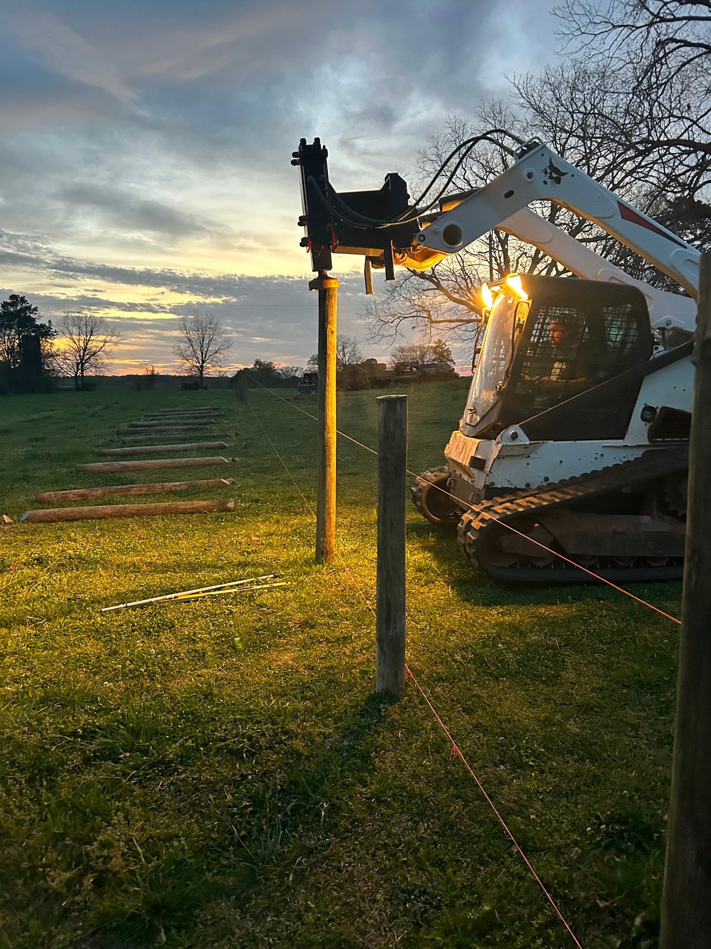 A skid steer uses an auger to set fence posts in a grassy field at dusk; golden light.