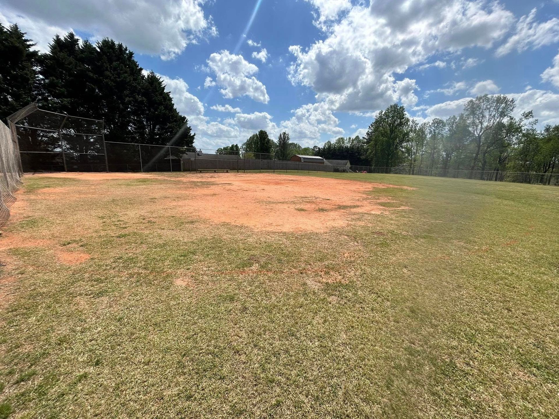 Grassy field with a large patch of reddish soil, under a cloudy blue sky.