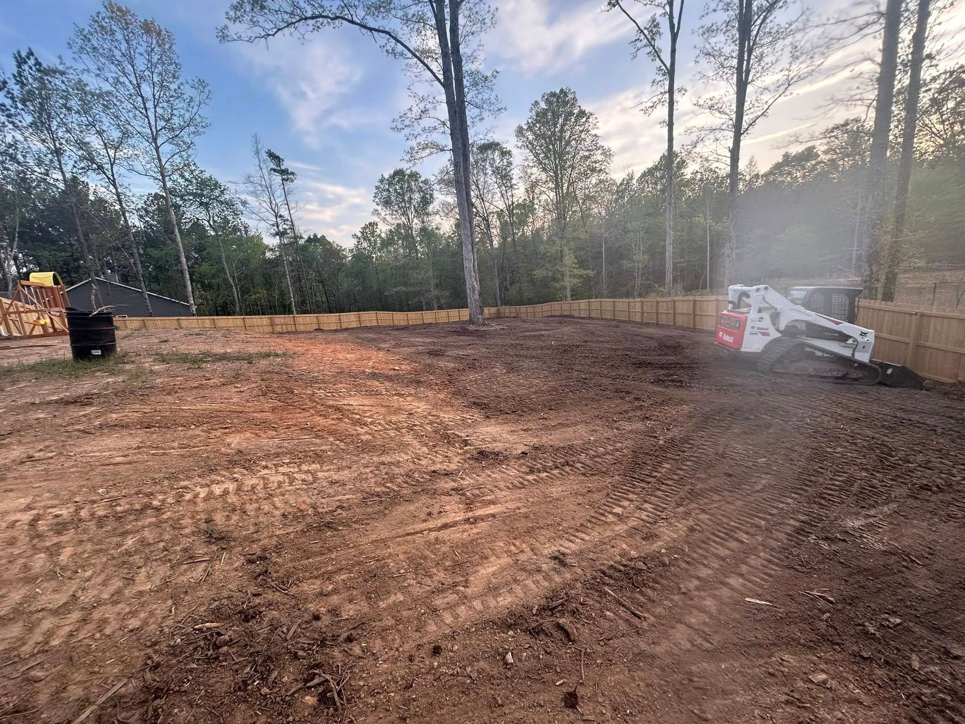 Bobcat working on a cleared dirt lot bordered by a wooden fence, trees in the background.