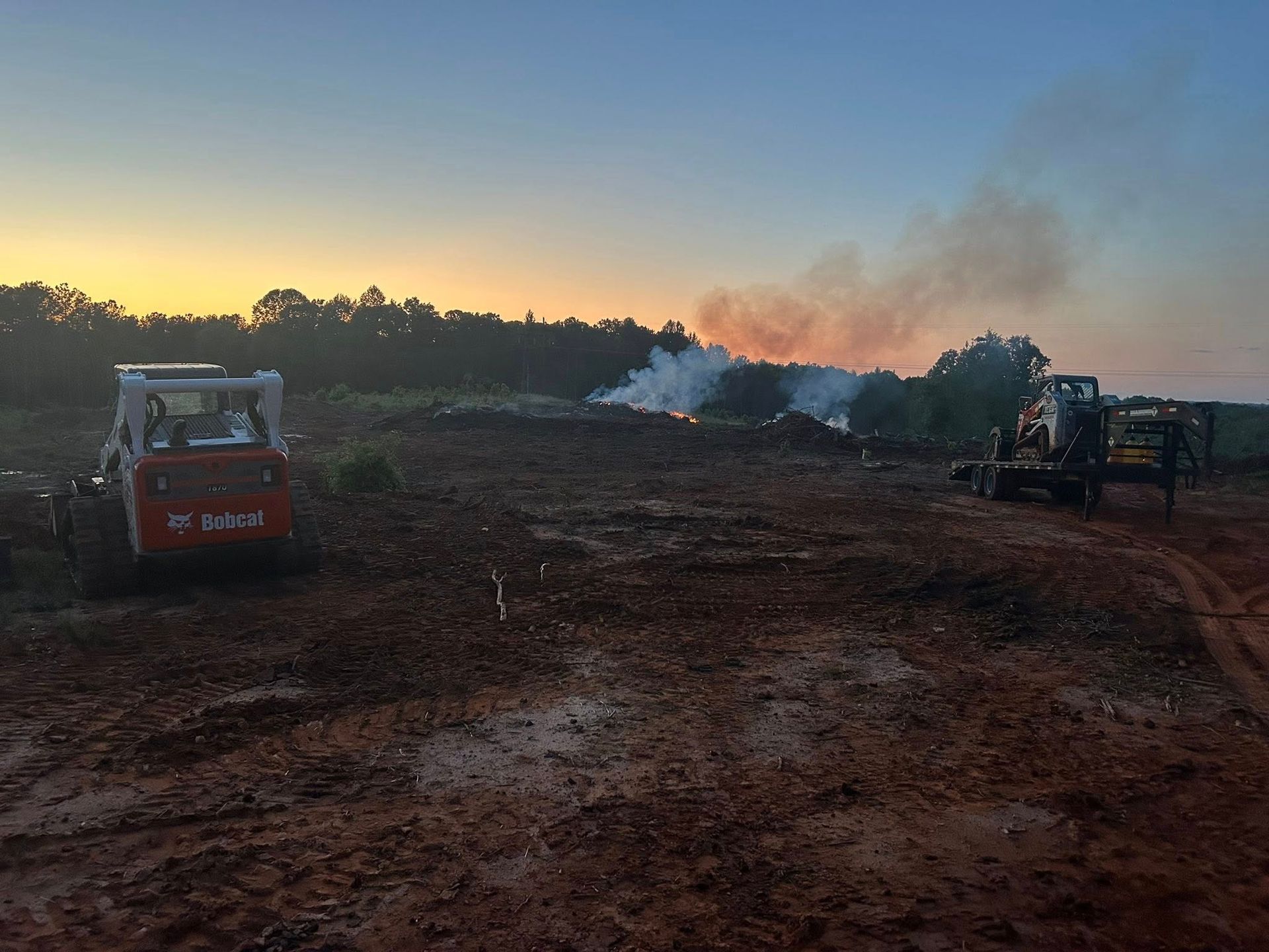 Construction site at dusk with smoking debris pile and two bobcat loaders.