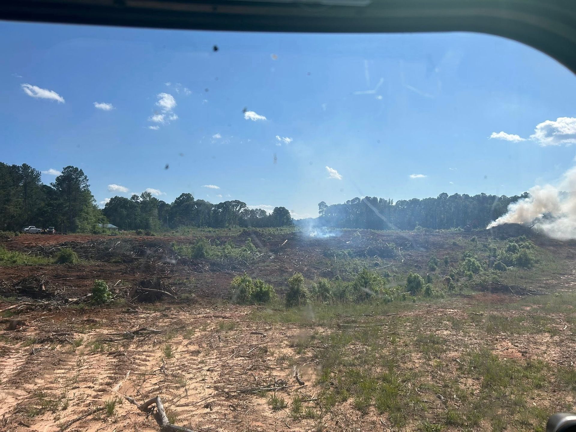 Field with smoke rising from a burning pile under a blue sky, trees in the background.