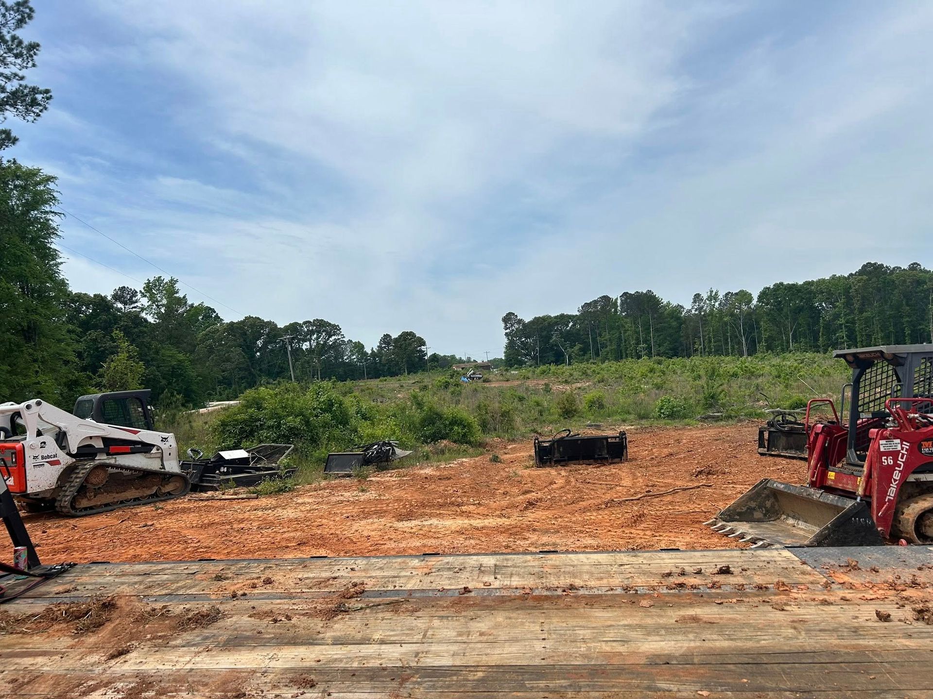 Construction site with red dirt, two small tractors, and treeline under a cloudy sky.