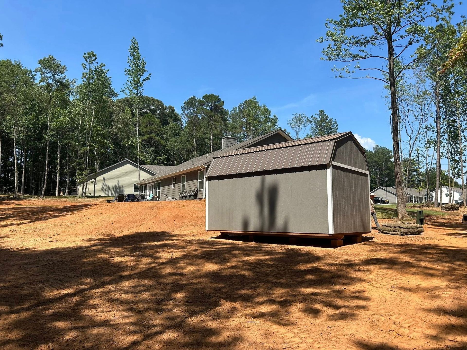 A grey storage shed in a cleared yard with a long building and trees in the background under a blue sky.