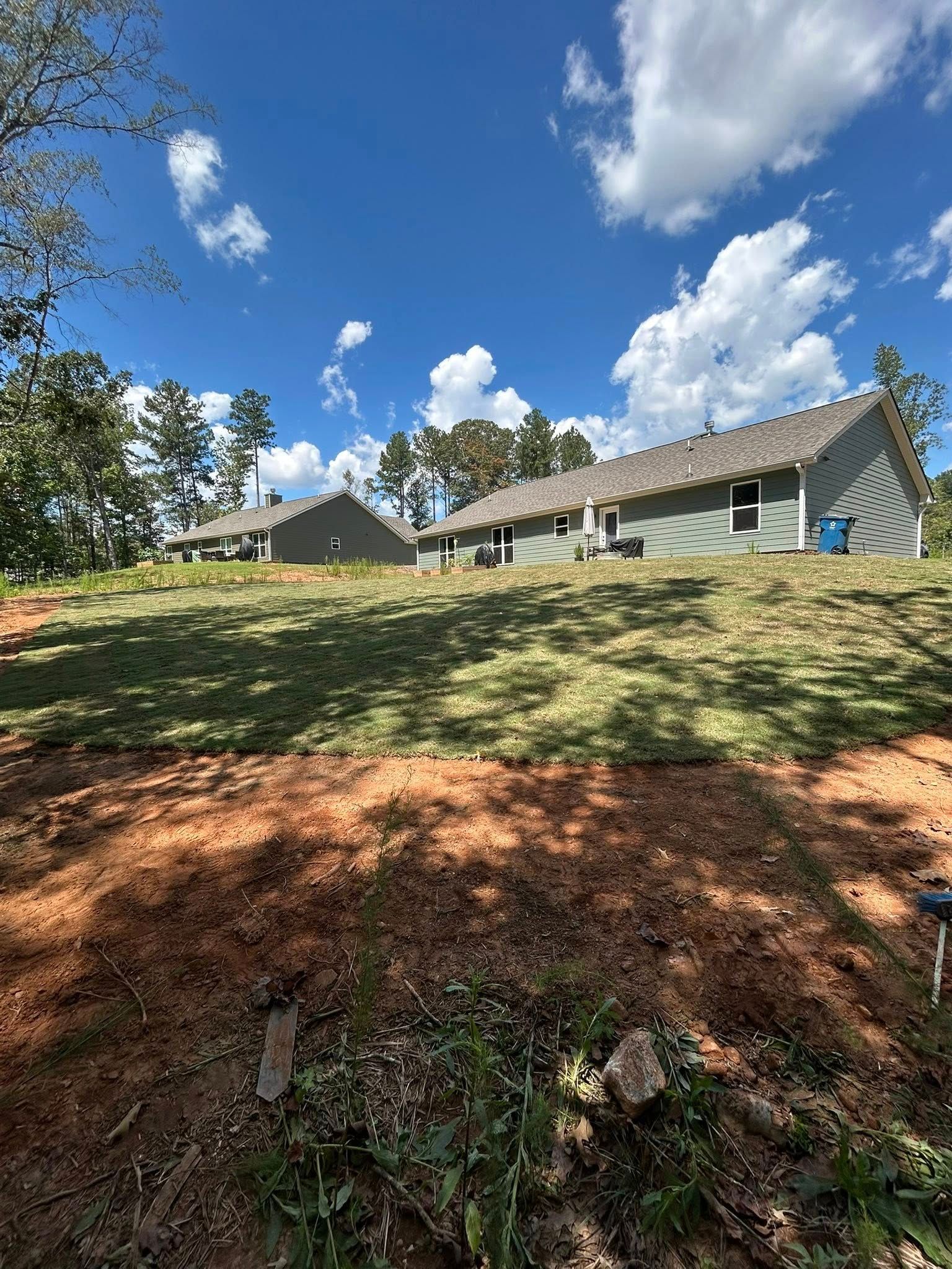 Two gray houses on a grassy hill under a blue sky with fluffy white clouds.