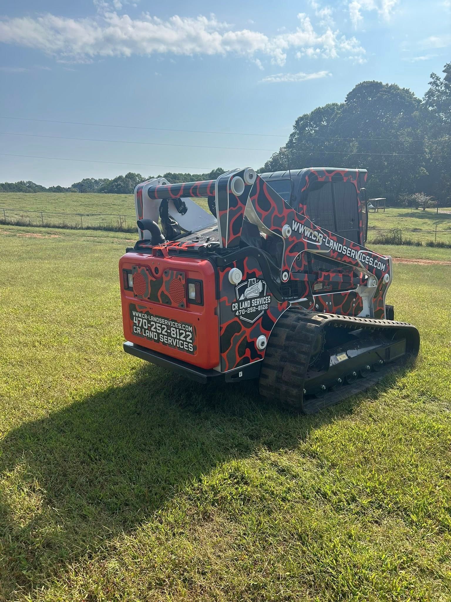 A red and black tracked robot on a grassy field with a tree-filled background on a sunny day.