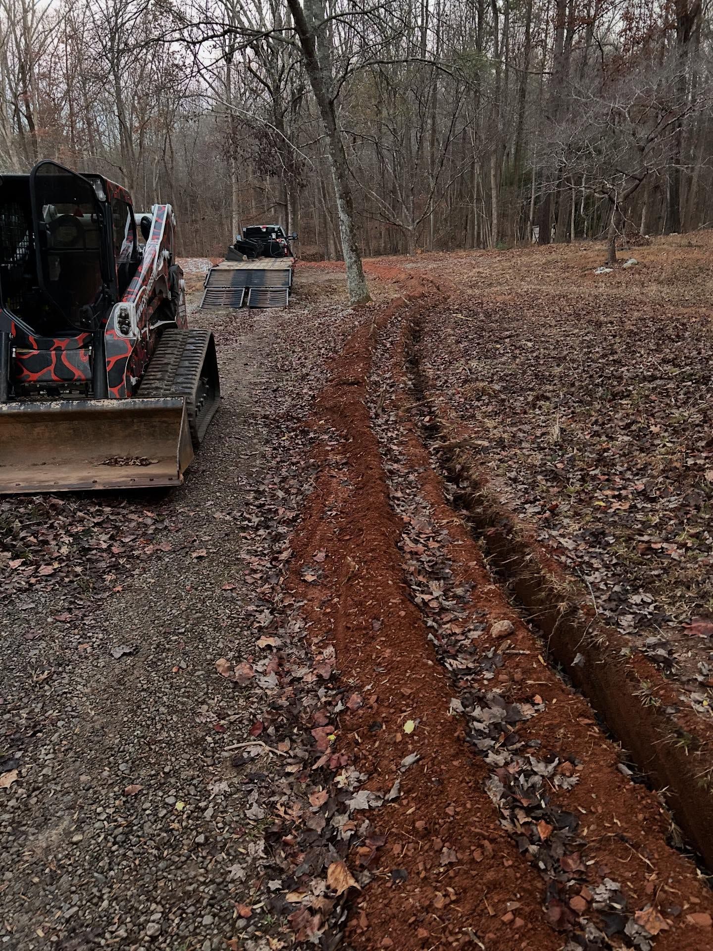A trenched path with red earth, a skid steer loader, and a second machine are in a wooded area.