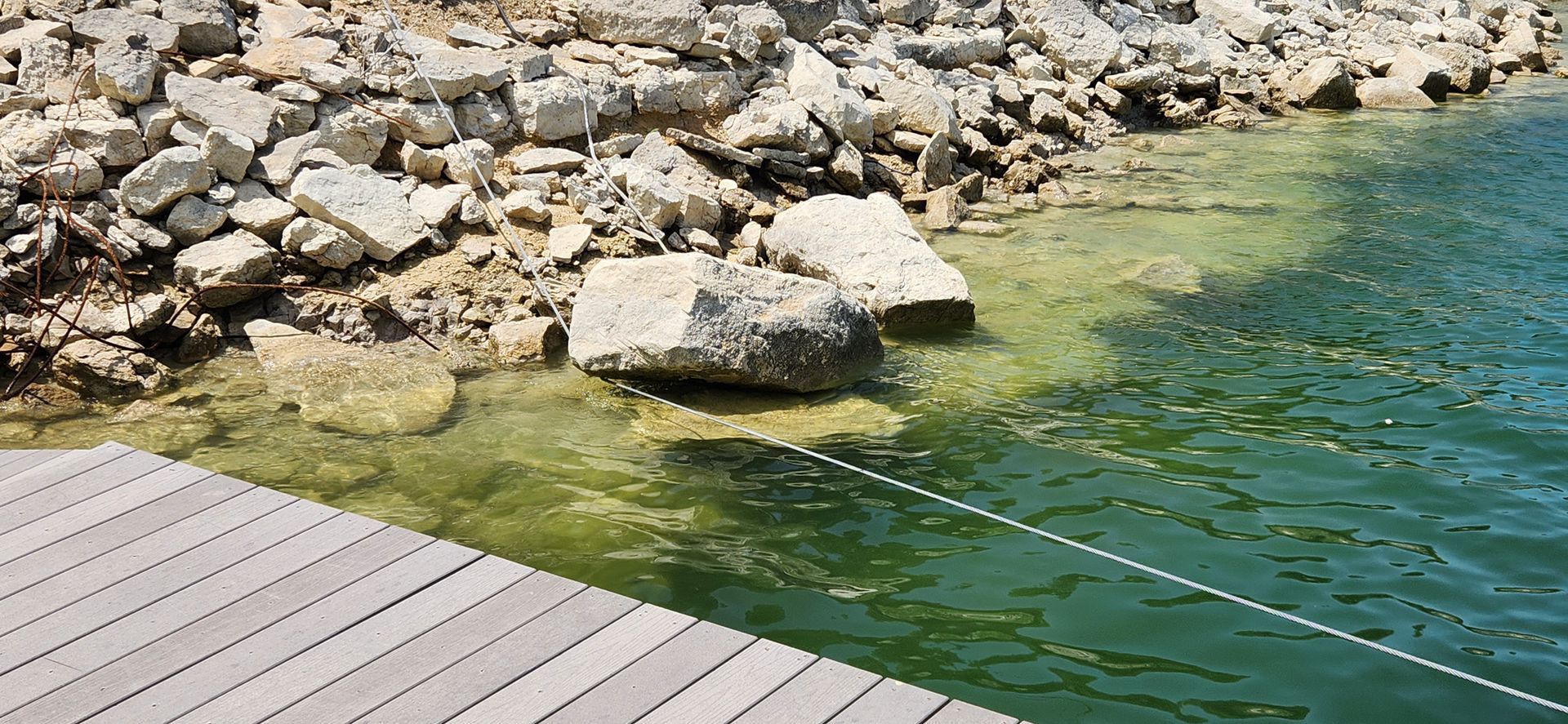 Wooden dock meeting green water with rocky shoreline in background.