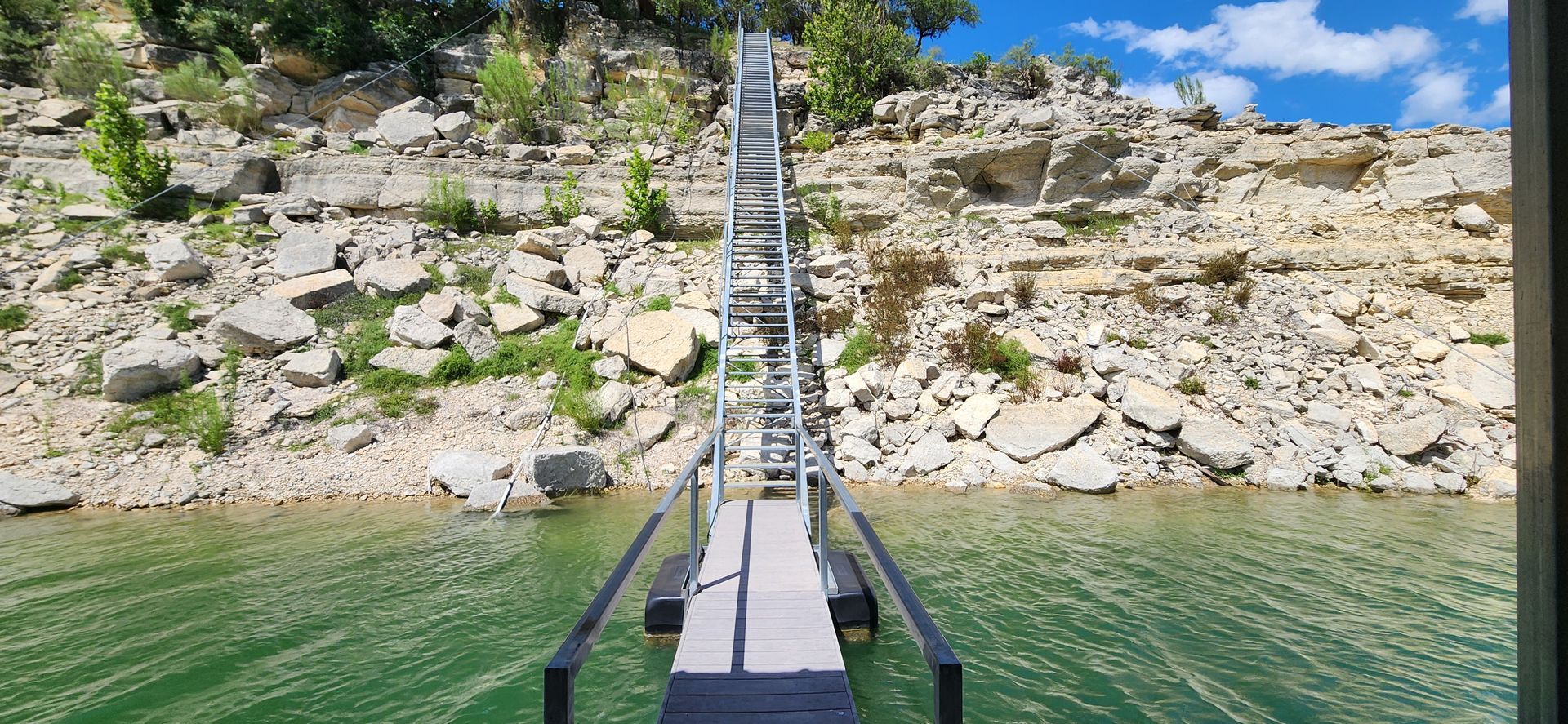 A long ladder extends from a dock into a lake, with a rocky hillside in the background under a blue sky.