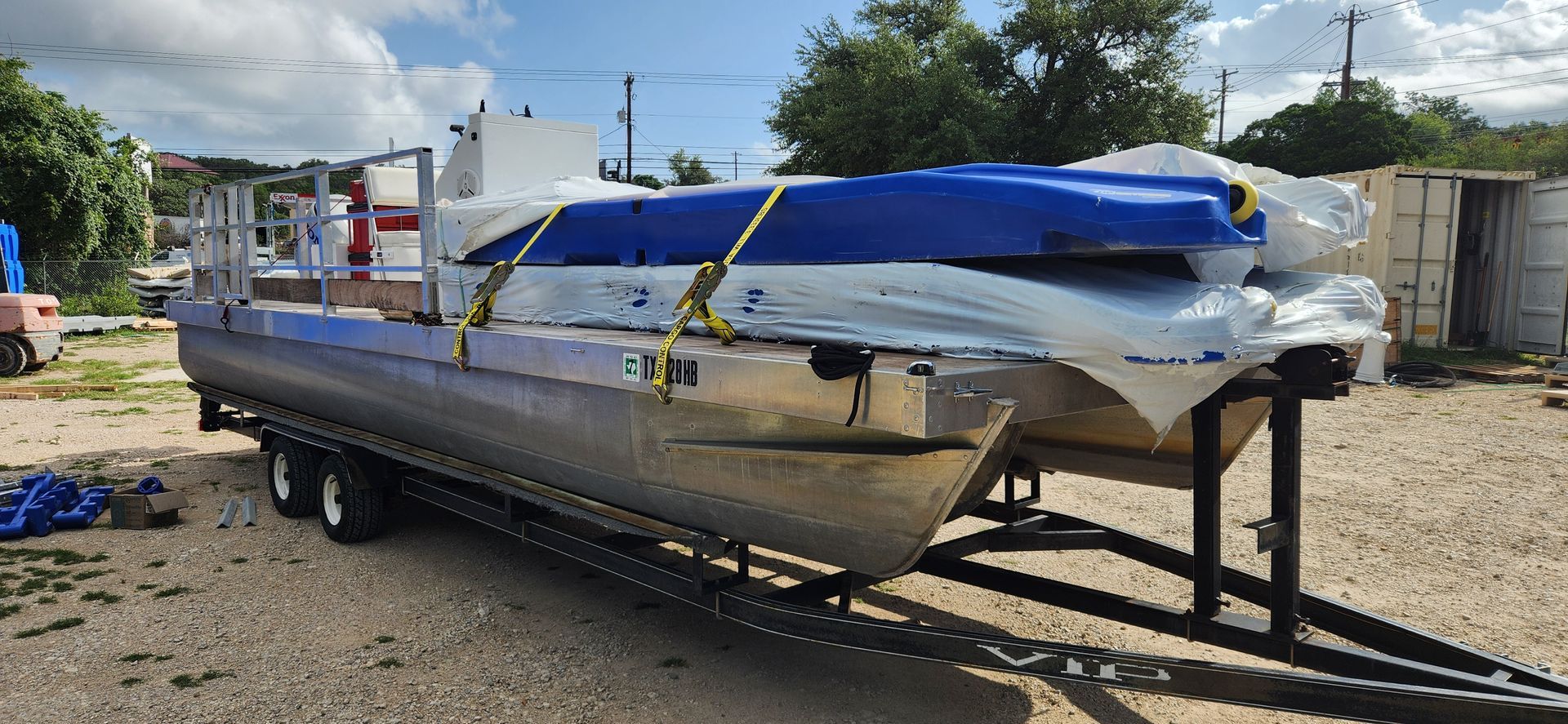 A pontoon boat on a trailer is covered in plastic and secured with straps outdoors.