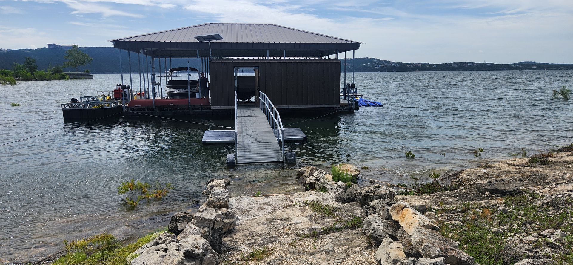 A dock with a covered boat slip on a lake, accessible by a ramp from a rocky shore.