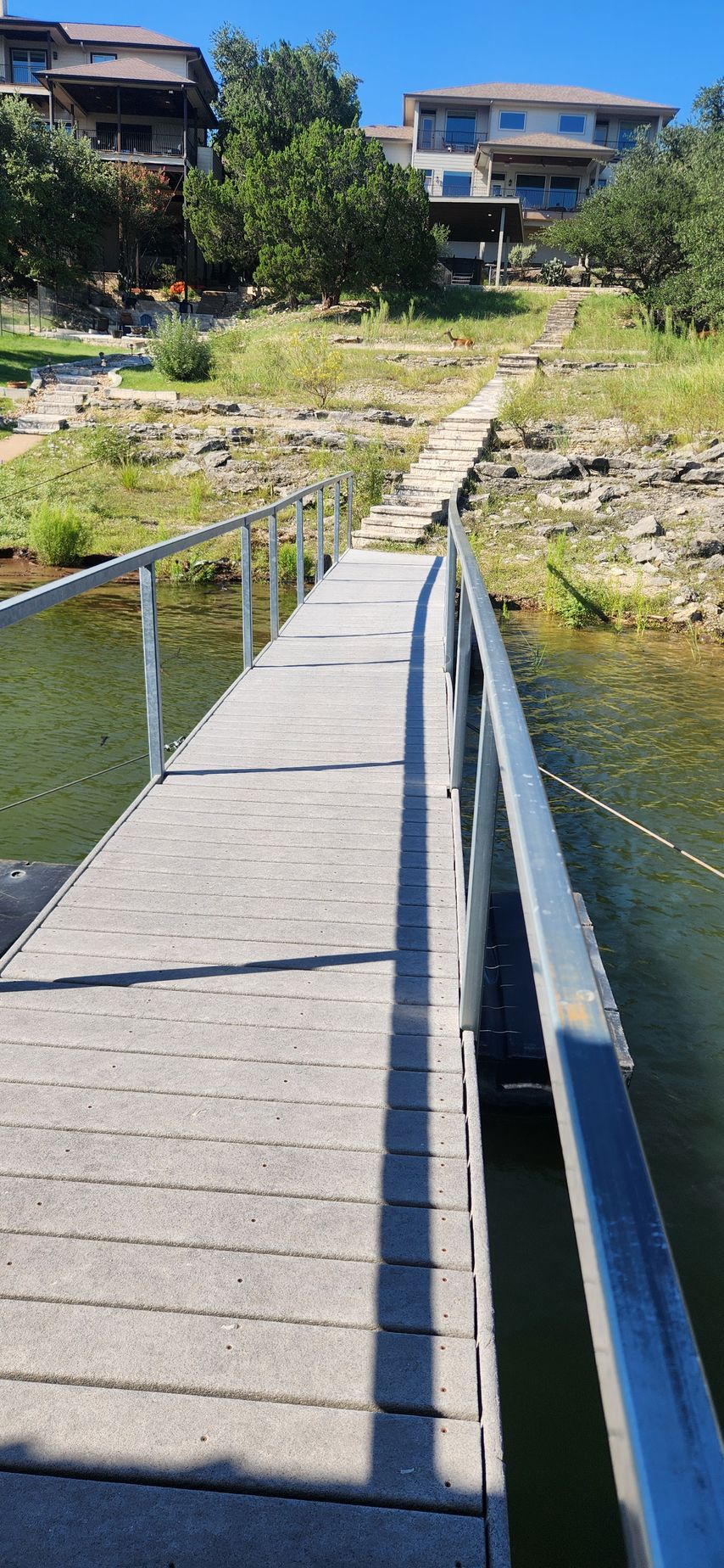 A wooden pier with metal railings stretches across water toward a rocky shoreline with houses.