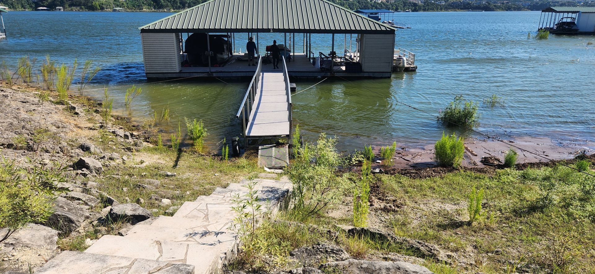 A dock leads to a floating structure on a lake. Greenery covers the shore on a sunny day.