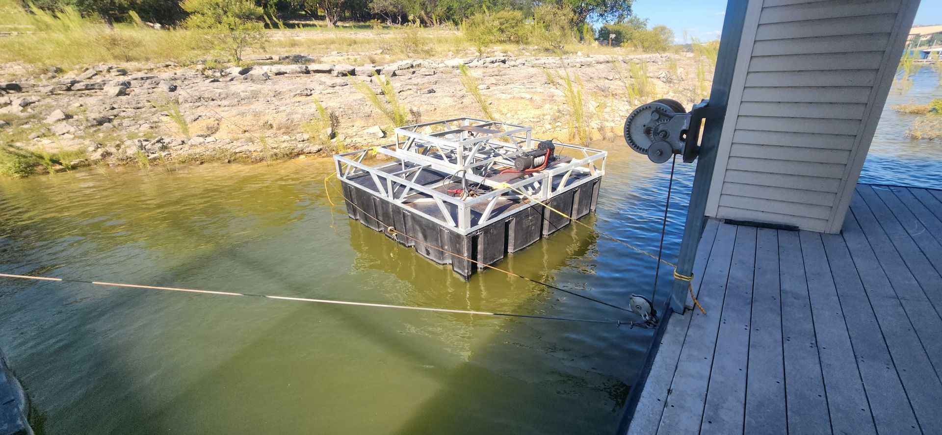 A large, floating cage in water, connected to a dock on the right, near a shoreline with trees in the background.