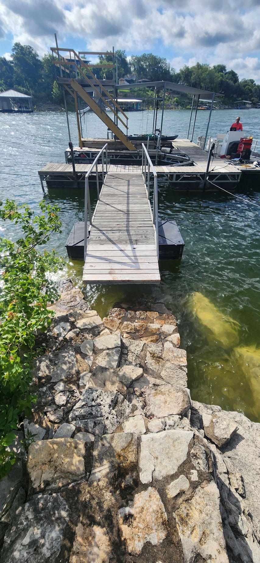 A wooden dock extending from a stone shore into a lake. The sky is cloudy.