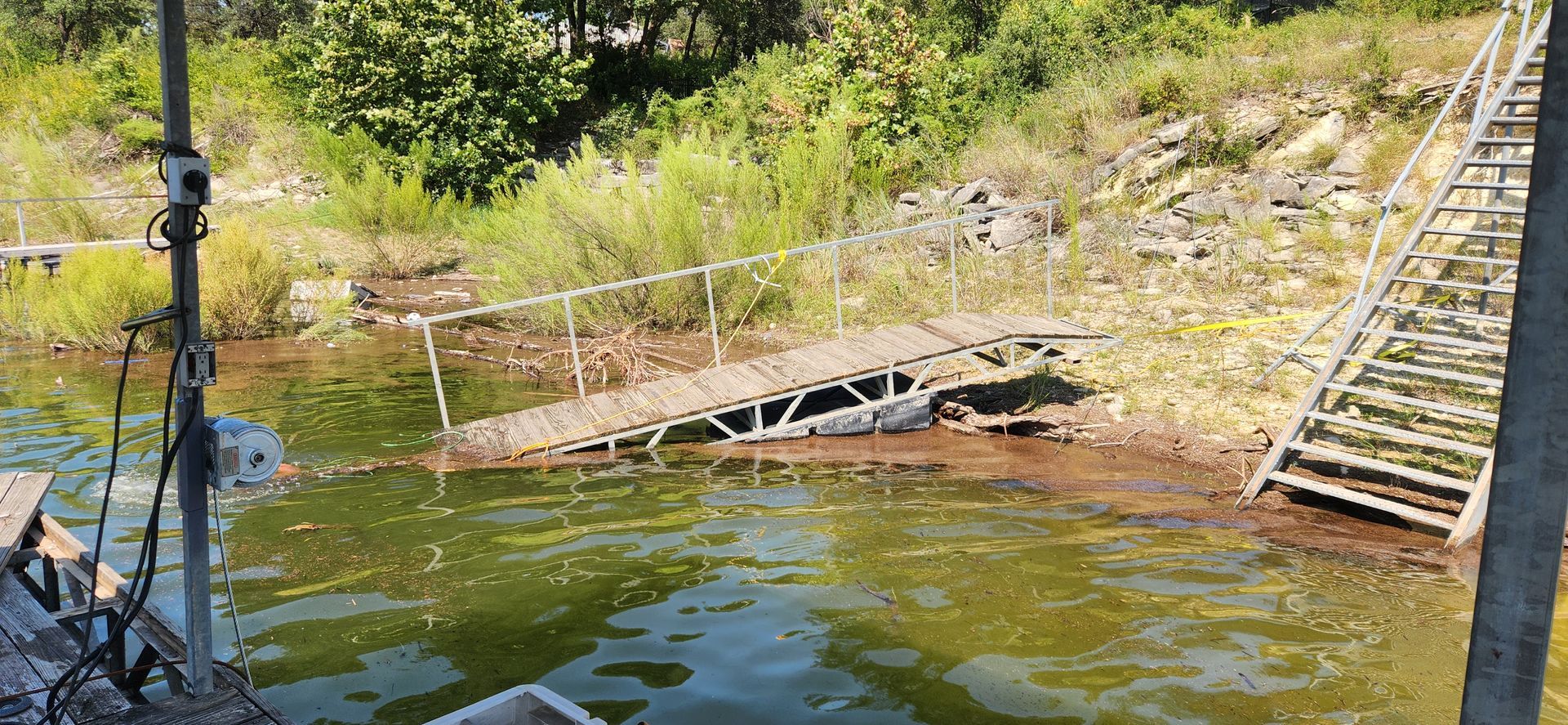 A dilapidated wooden dock slopes into murky water, next to stairs leading up a grassy bank.