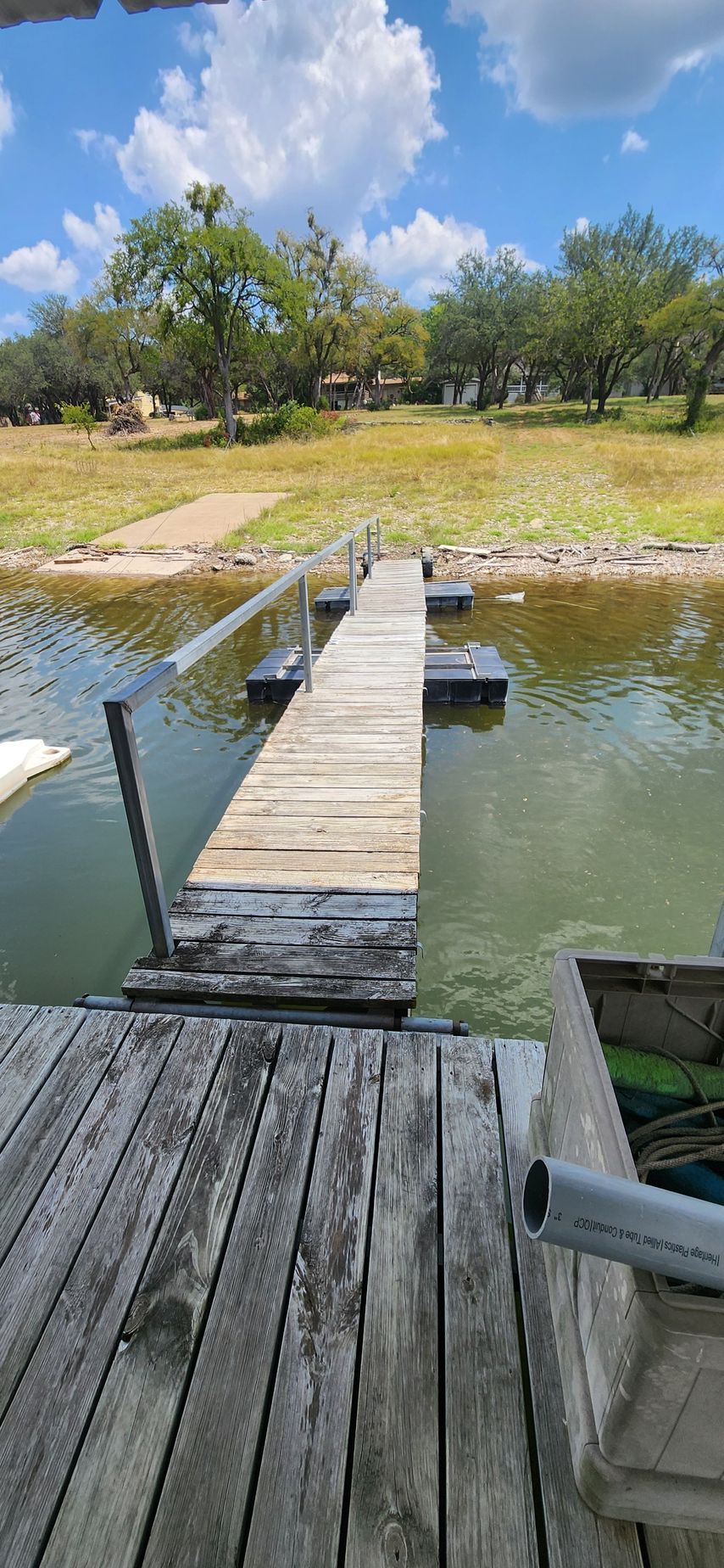 Wooden dock extending over water towards a shoreline with trees, under a partly cloudy sky.