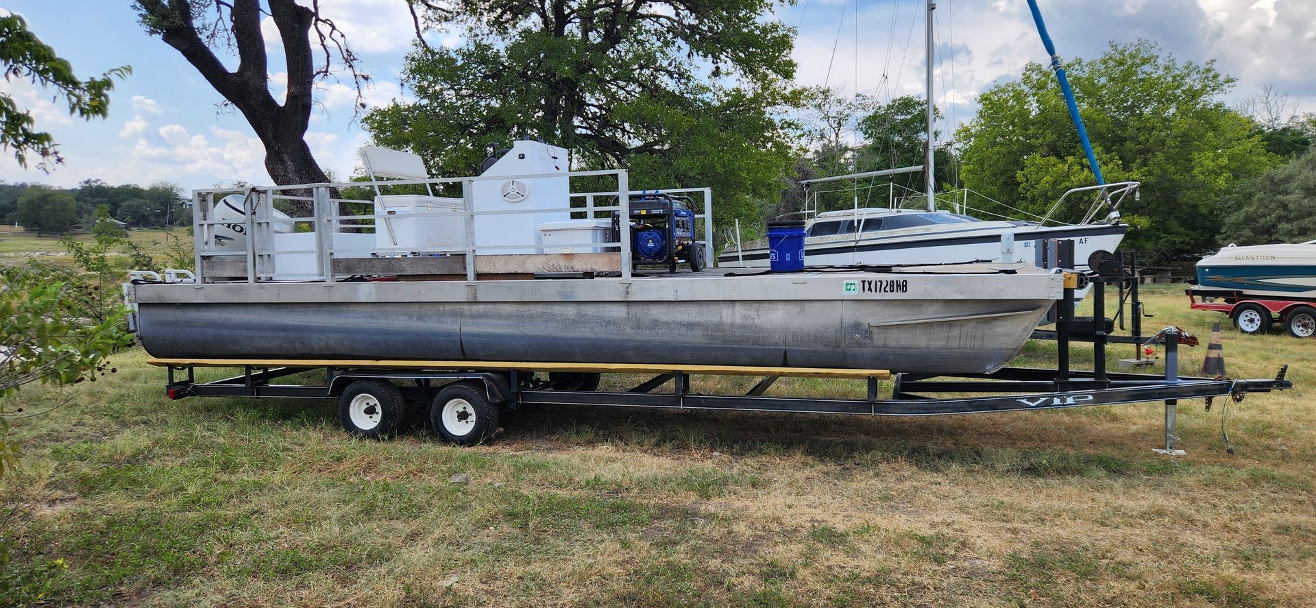 A large pontoon boat on a trailer, parked on grass, with trees in the background.