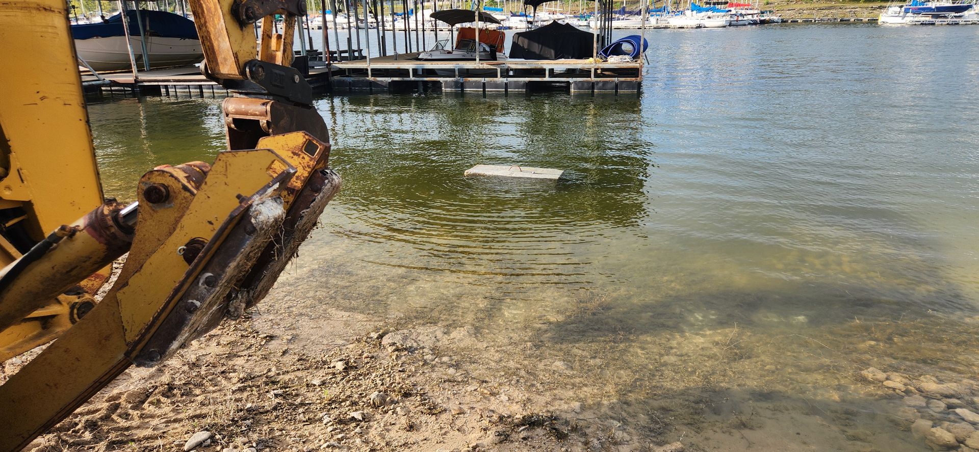 A yellow excavator arm near a muddy shore, facing a dock and water with boats.