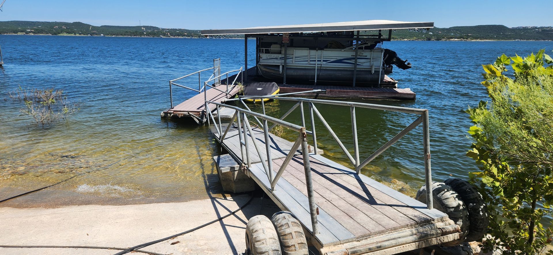 A floating dock with a covered structure on a sunny lake, with a ramp leading to the shore.