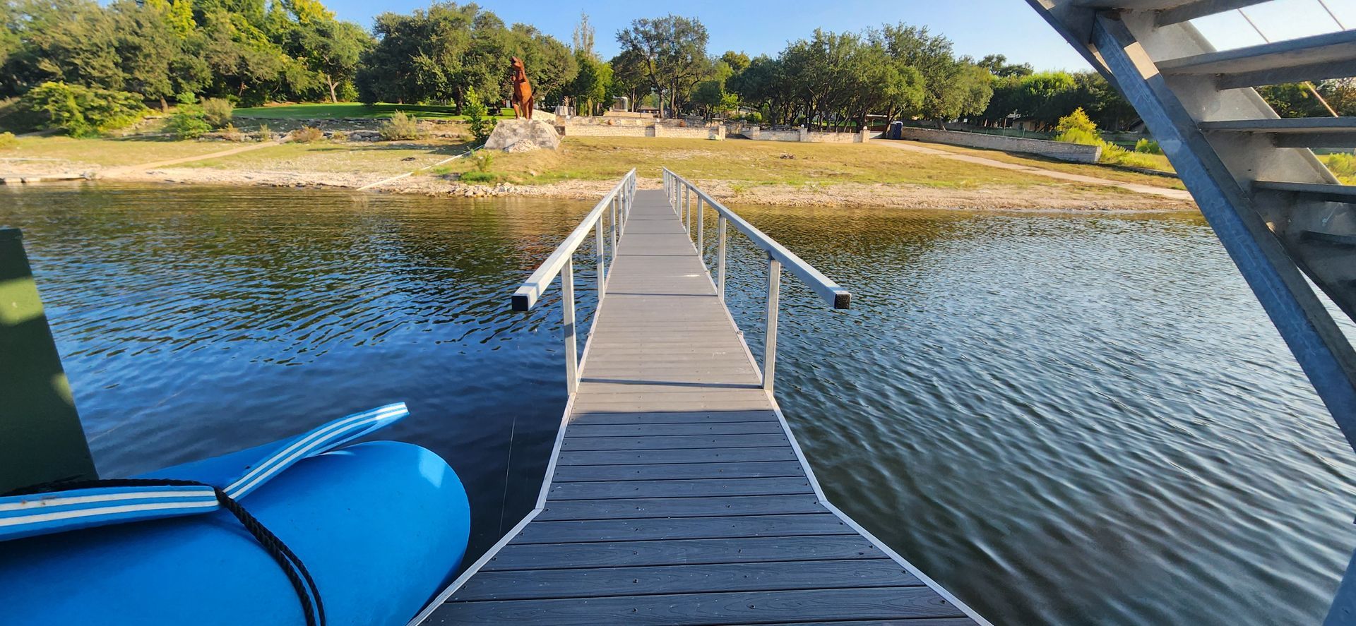 A dock extends into a body of water. Trees line the opposite shore. A blue inflatable boat is on the left.