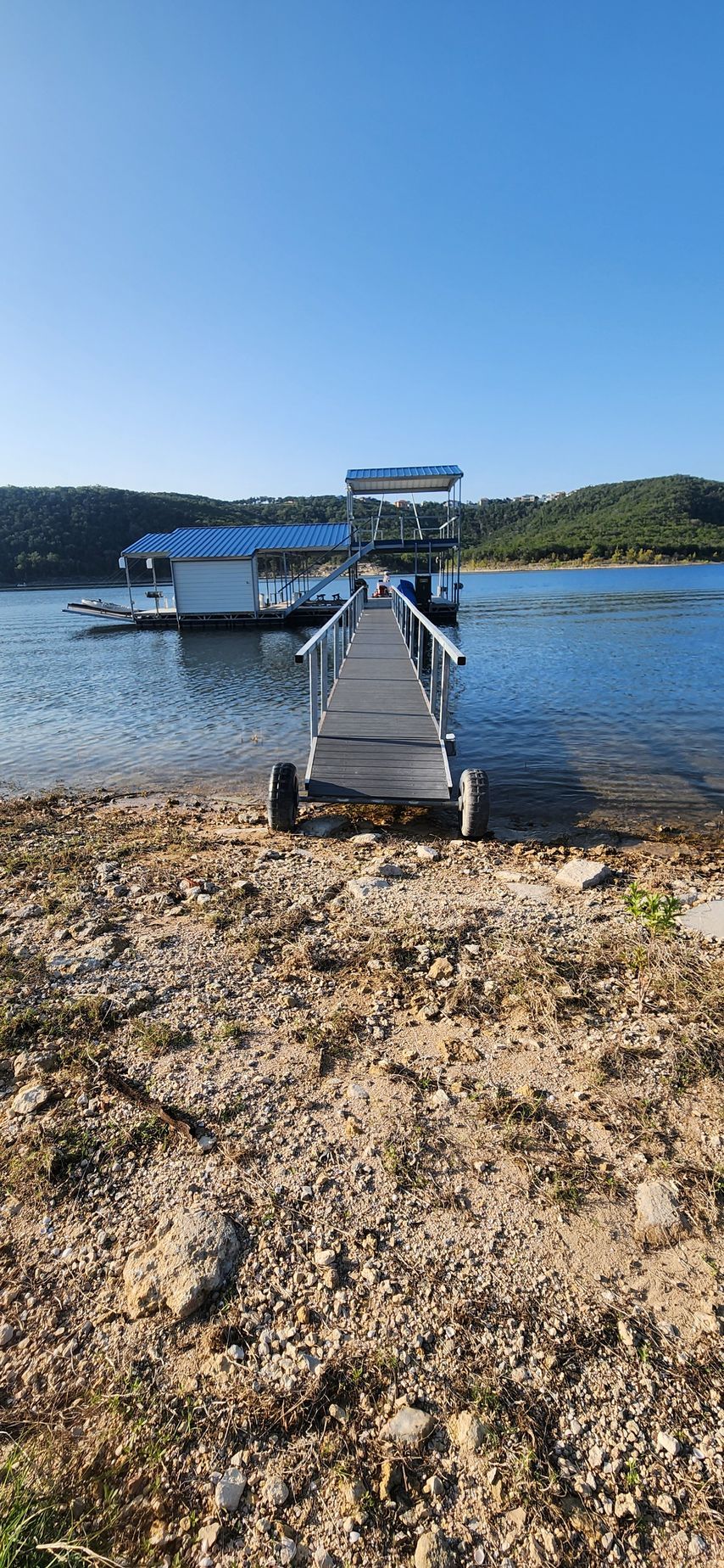 A wooden pier leads to a floating house on a lake, clear blue sky overhead.
