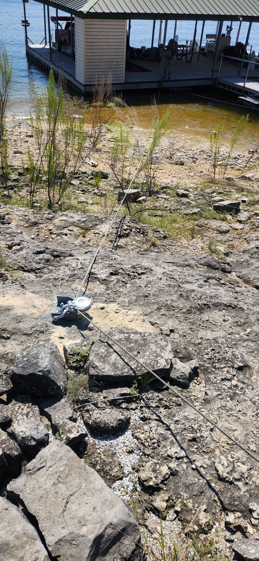 A white drone on a rocky shore by a dock, green water, and small bushes in sunlight.