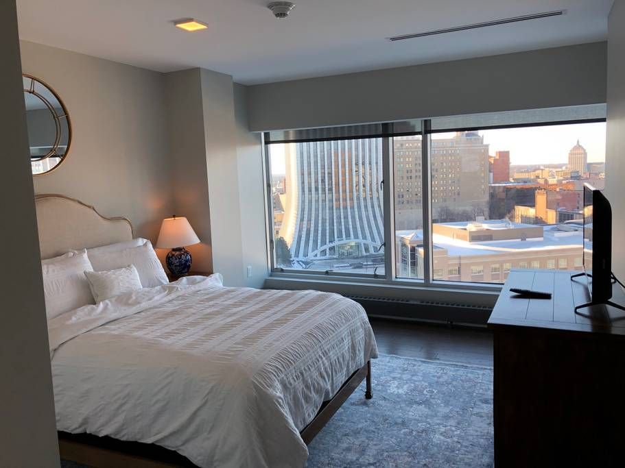 Bedroom with bed, city view from large window, TV on dresser, mirror on wall.