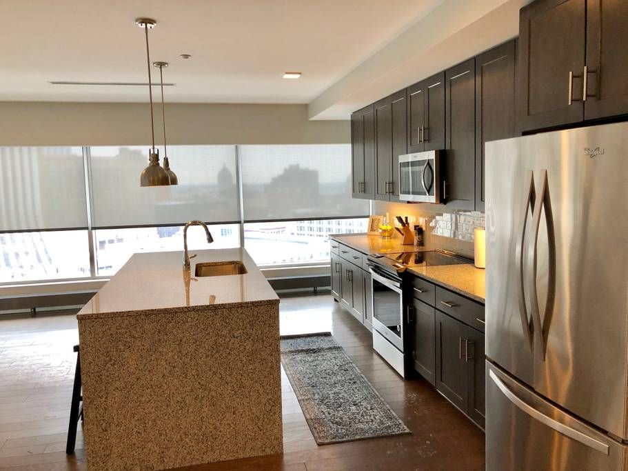 Modern kitchen with stainless steel appliances, island, and dark cabinets. Large windows overlook a city.