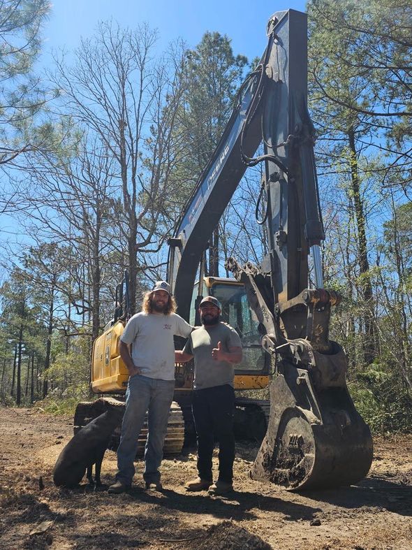 Two men stand in front of a large excavator in a clearing, one giving a thumbs up. The setting is a wooded area on a sunny day.
