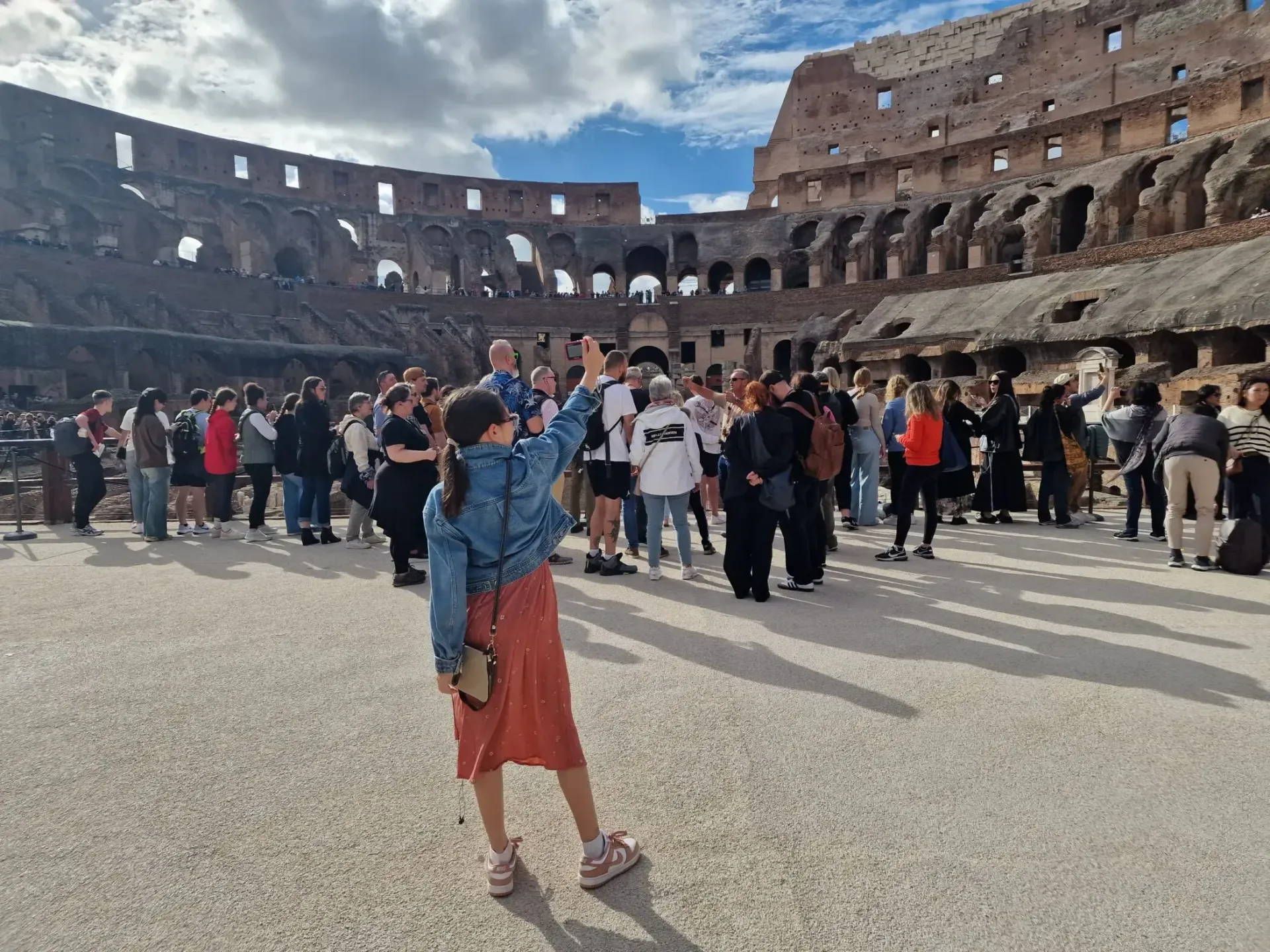 Person waving inside the Colosseum in Rome, surrounded by tourists, sunny day.