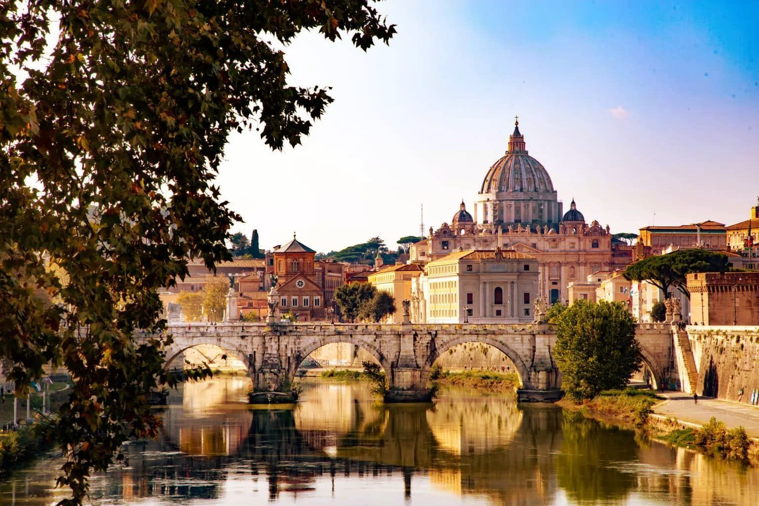 River view of Rome with bridge, buildings, and St. Peter's Basilica dome under blue sky.