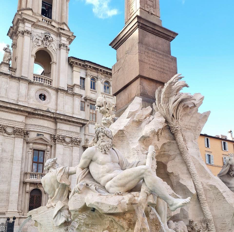 Sculpture with a reclining figure, obelisk, and church in a sunny plaza.