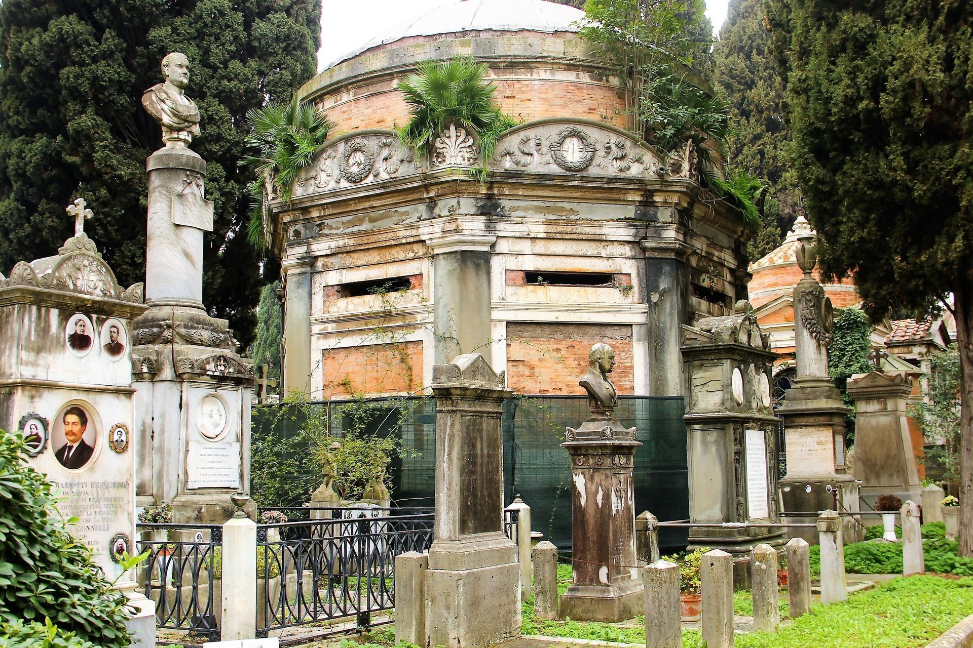 Ancient, weathered cemetery with ornate tombs and mausoleum. Overgrown foliage with a cloudy sky.