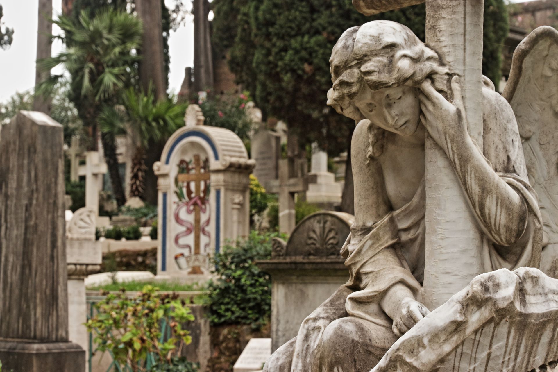Stone angel statue leaning on a cross in a cemetery, surrounded by headstones and trees.