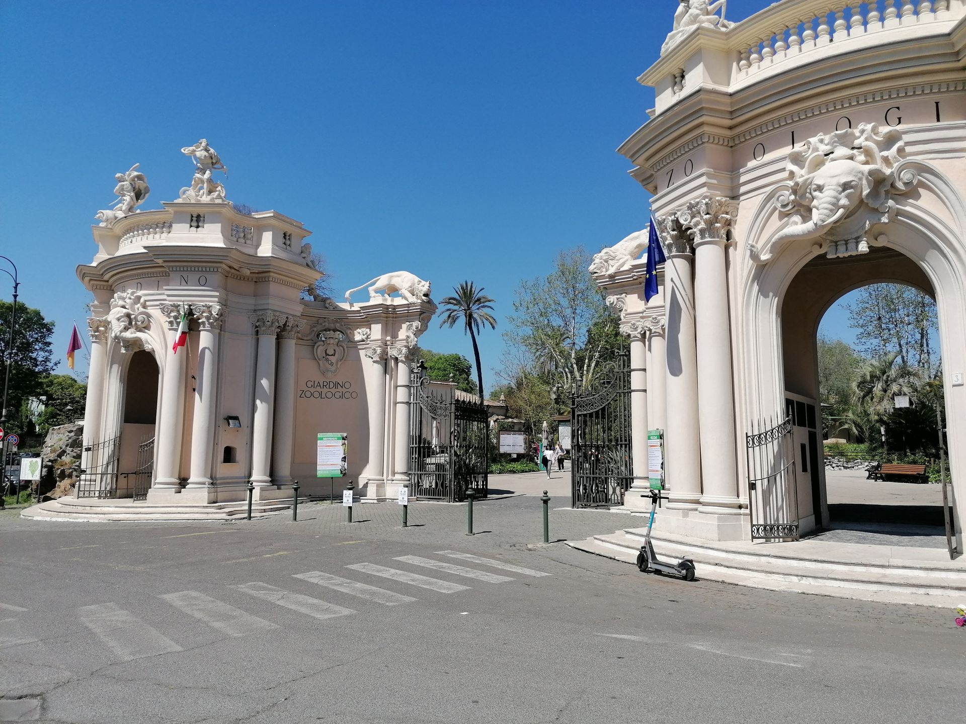 Ornate archway entrance to a park on a sunny day. Two columns support sculpted details and flagpoles.