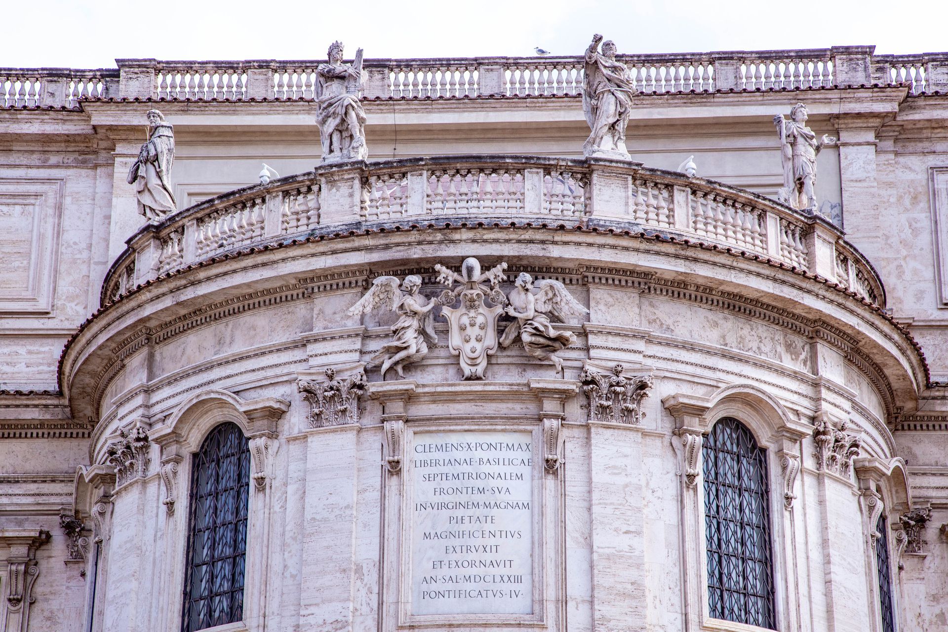 Stone bridge with statues over a river in Rome, Italy. Buildings and trees line the banks under a cloudy sky.