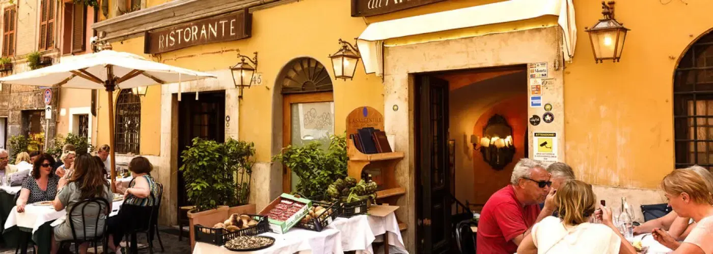 Restaurant with outdoor seating. People dine at tables beneath an umbrella and awning on a sunny day.
