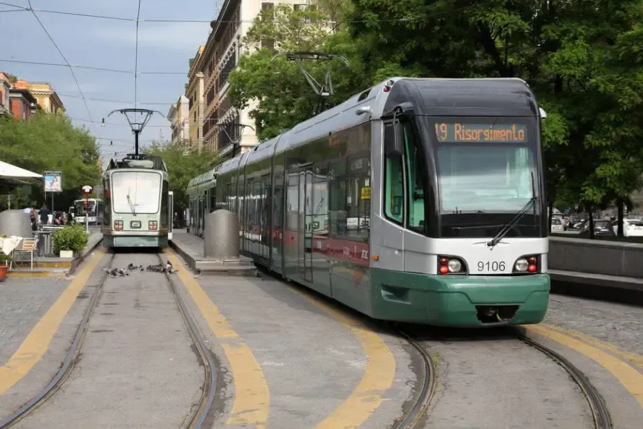 Two trams on tracks at a station. Modern tram is silver, black, and green. Old tram is white and green. City setting.