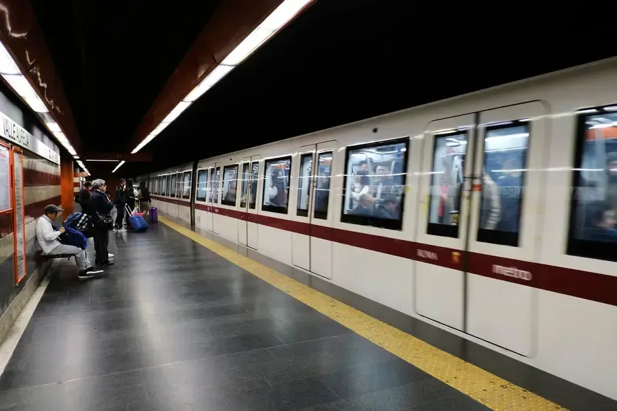 Subway train pulls into station. People wait on platform; dark ceiling, white train, red trim.