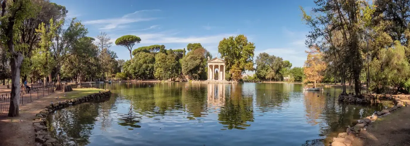 A calm lake reflects trees and a small temple in a park under a blue sky.