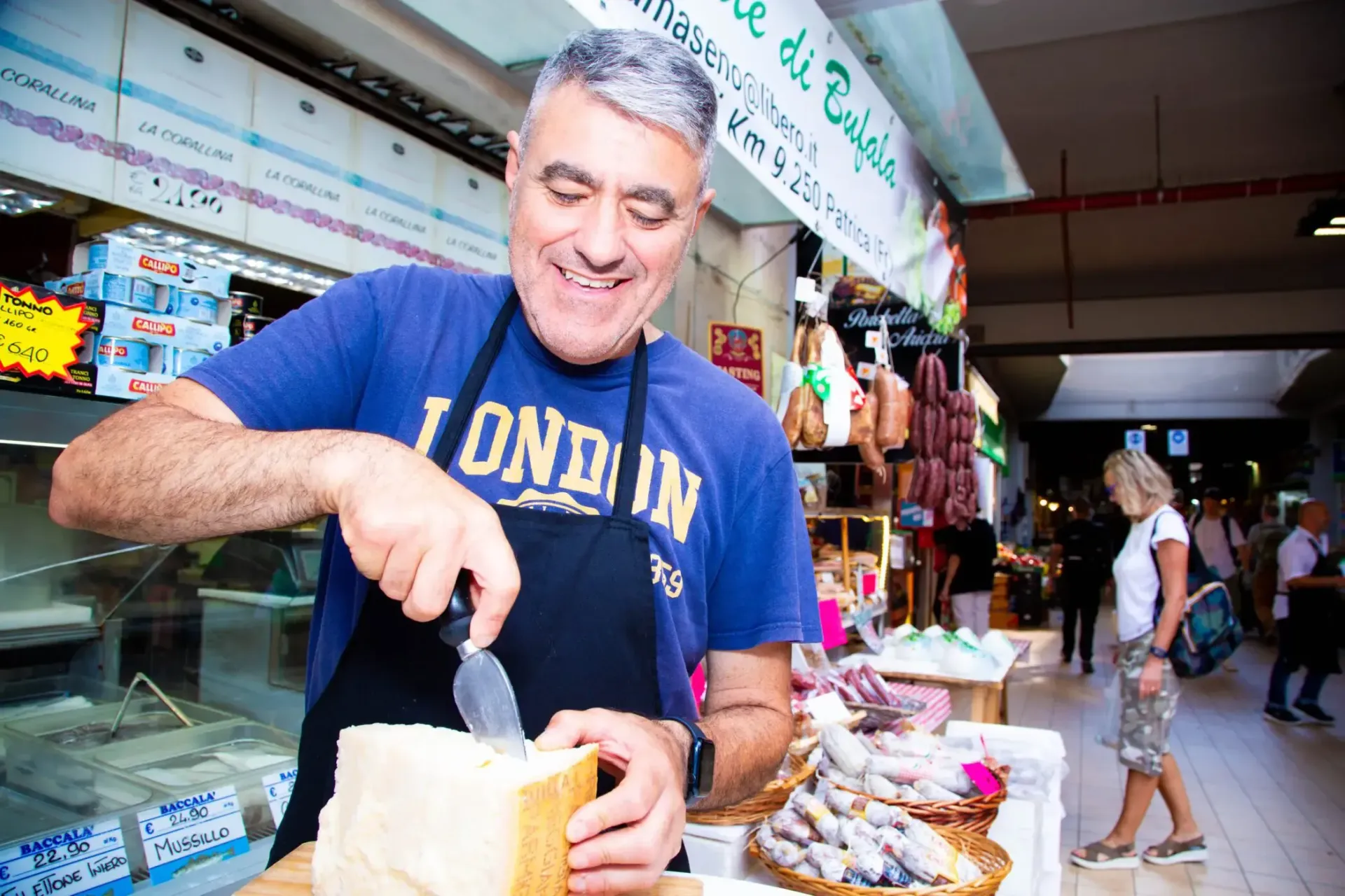 Man cutting cheese at a market stall, smiling, wearing a London shirt and apron.
