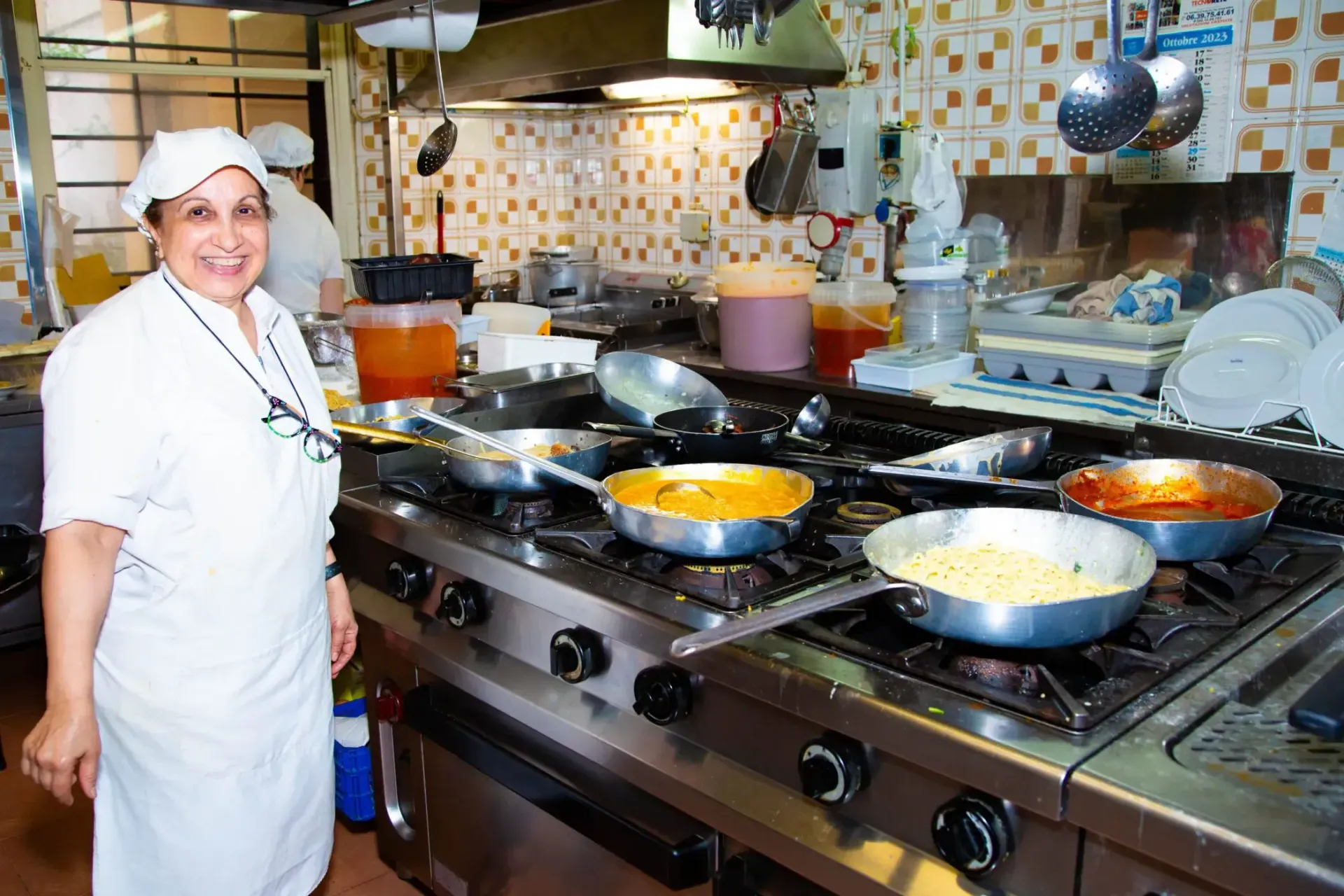 Woman in chef's uniform smiles, standing in a commercial kitchen with pots on stove.