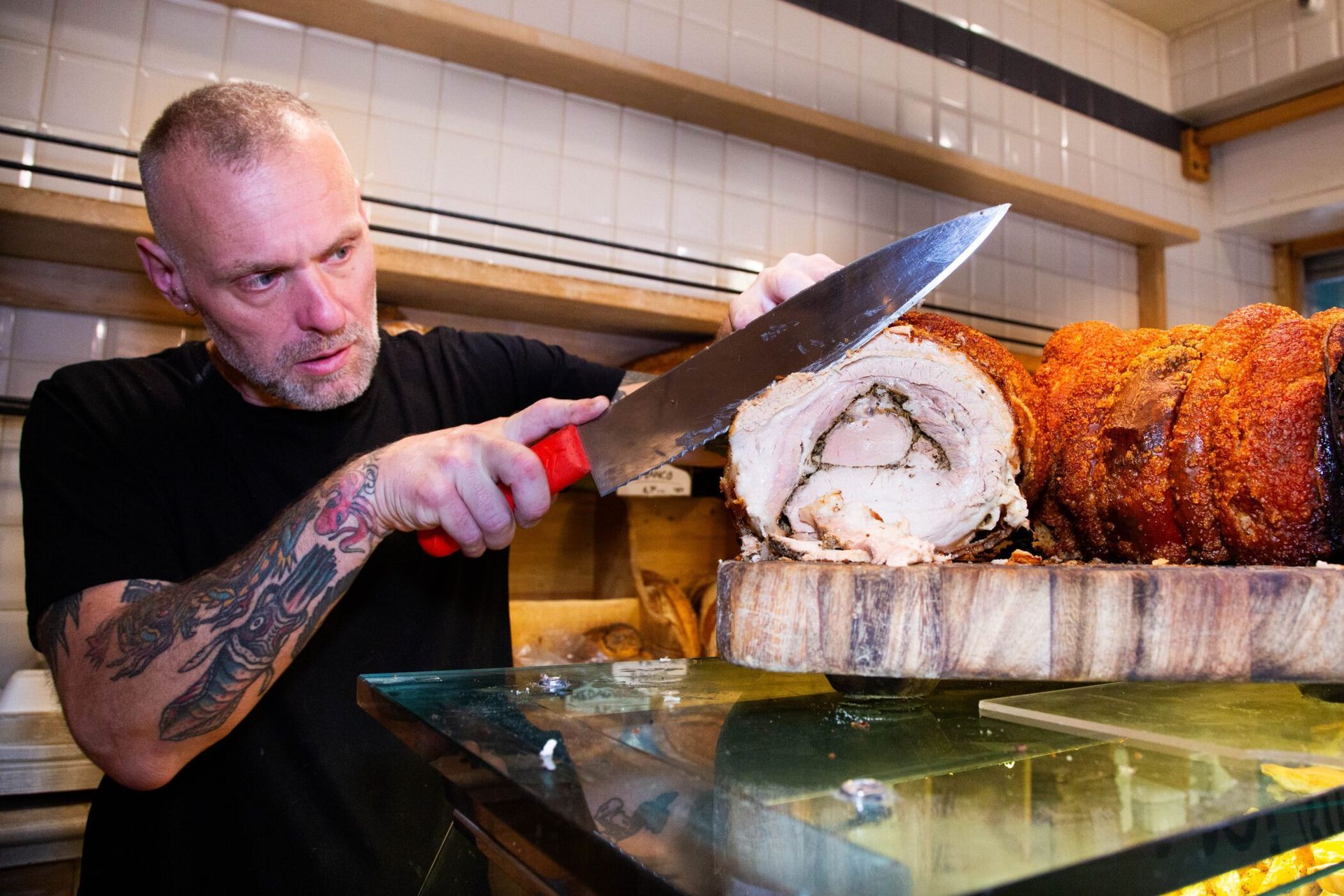 Man slicing roasted pork, likely in a restaurant. He has tattoos and focused expression, holds large knife.