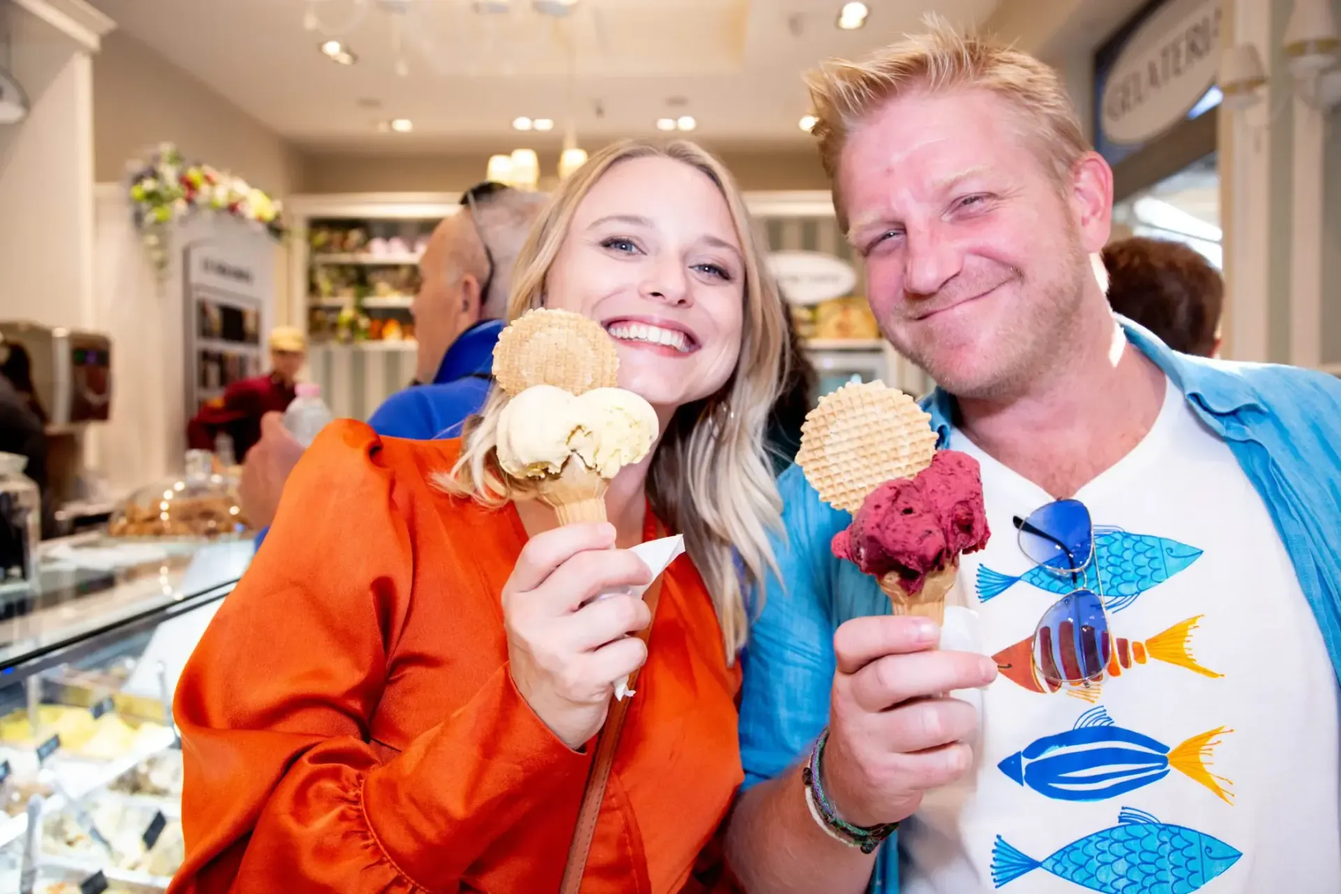 Couple smiling, holding ice cream cones in a shop. One wears orange, the other a fish-print shirt and blue shirt.